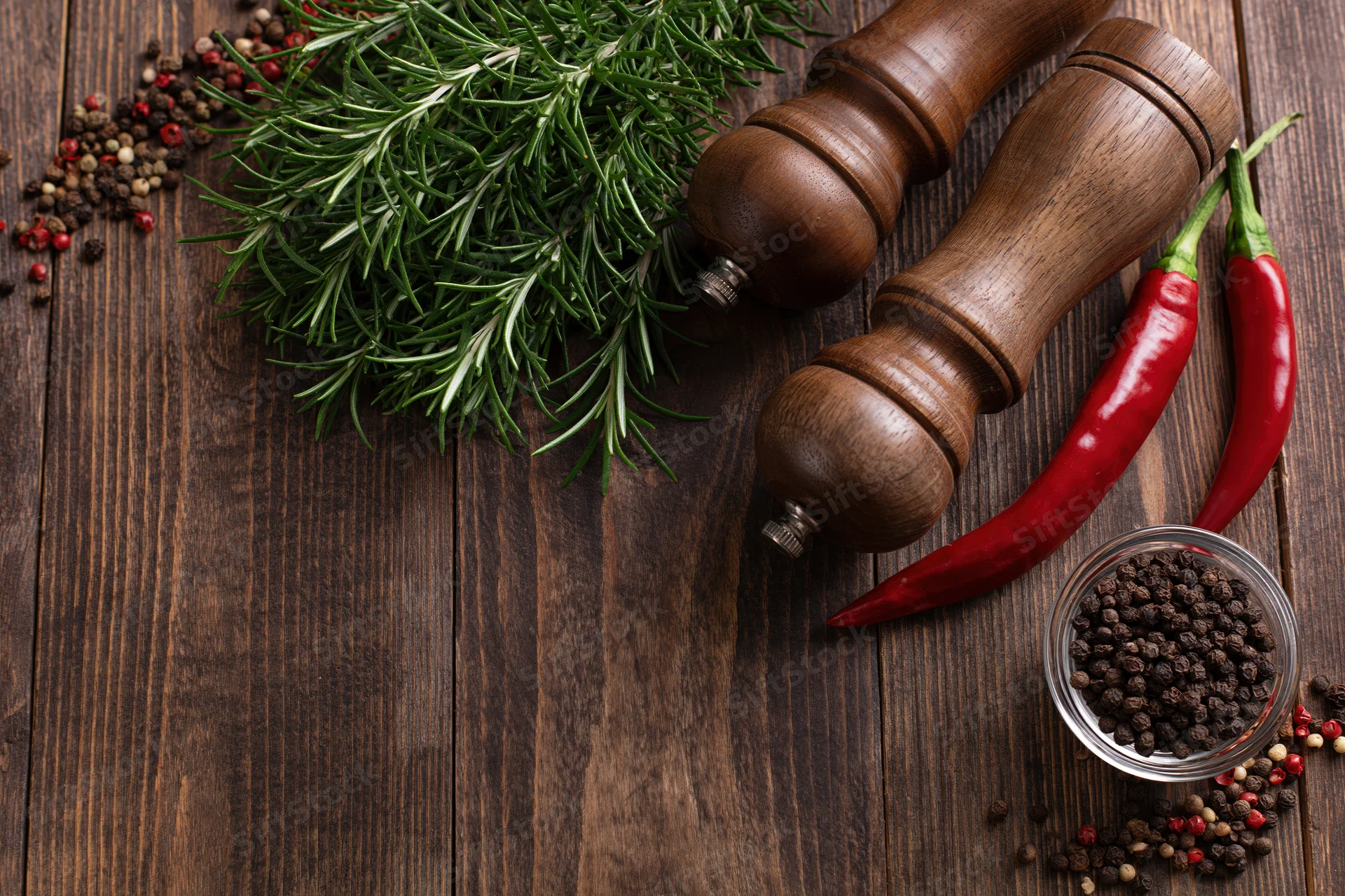 Fresh rosemary sprigs and red chili peppers with wooden spice grinders arranged on a dark wooden table in a flat lay composition