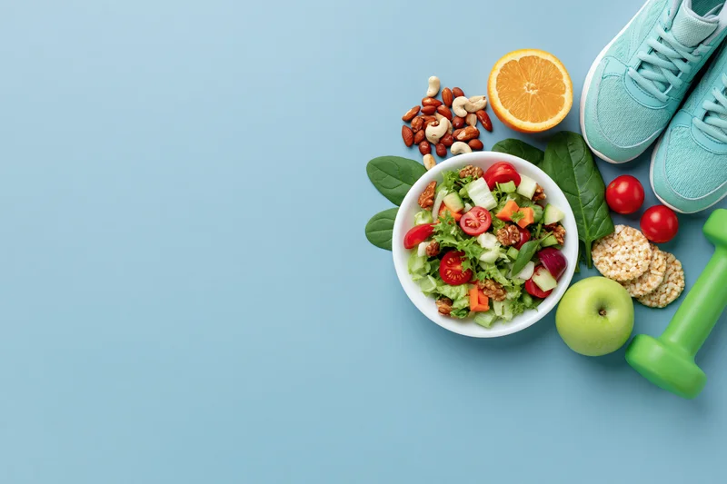 Salad with vegetables and fruit, nuts, apple, orange, sneakers and dumbbell arranged on blue background