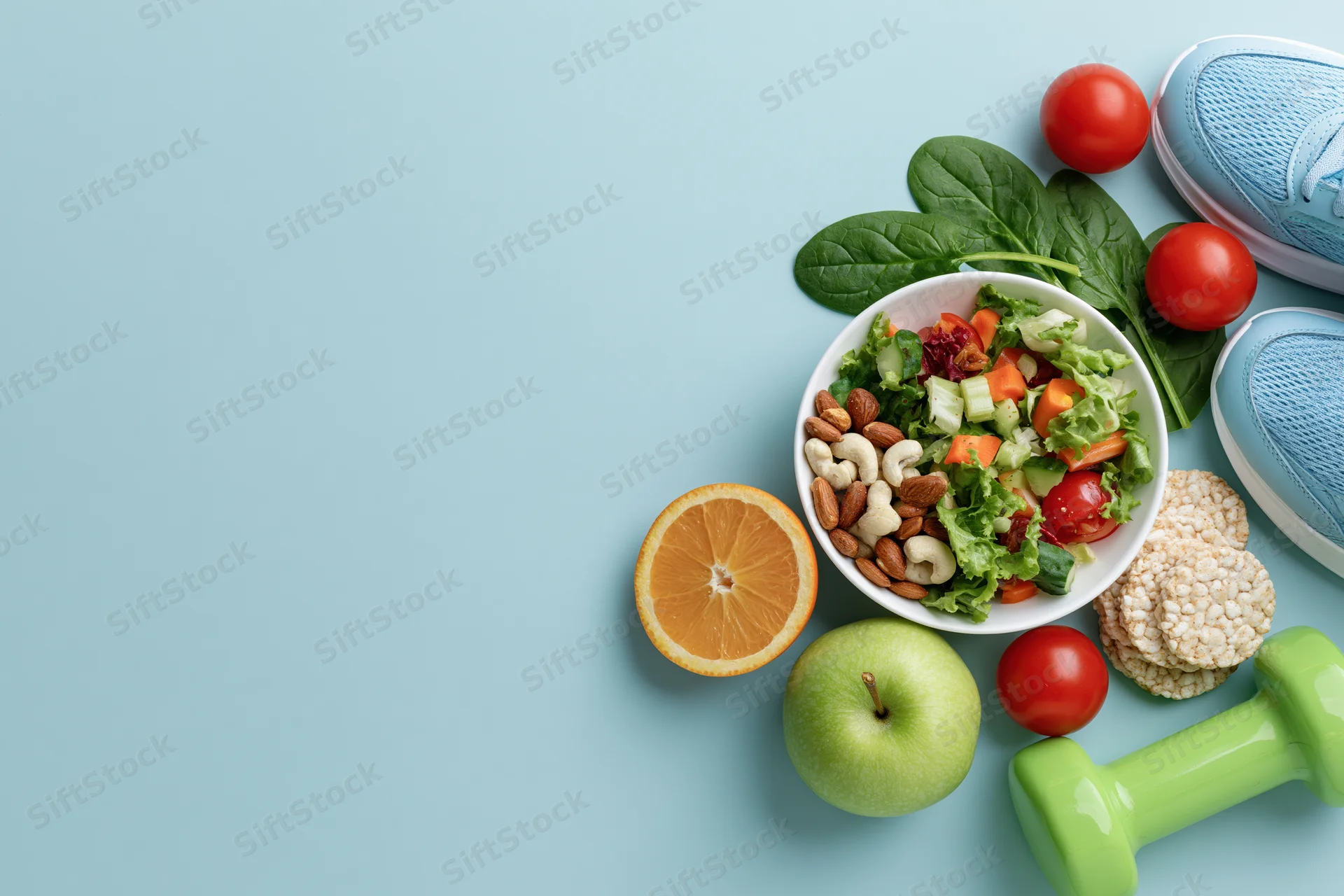 Salad with vegetables and nuts in a bowl surrounded by fruit, sneakers, and a dumbbell on a blue surface