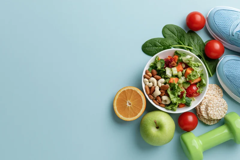 Salad with vegetables and nuts in a bowl surrounded by fruit, sneakers, and a dumbbell on a blue surface