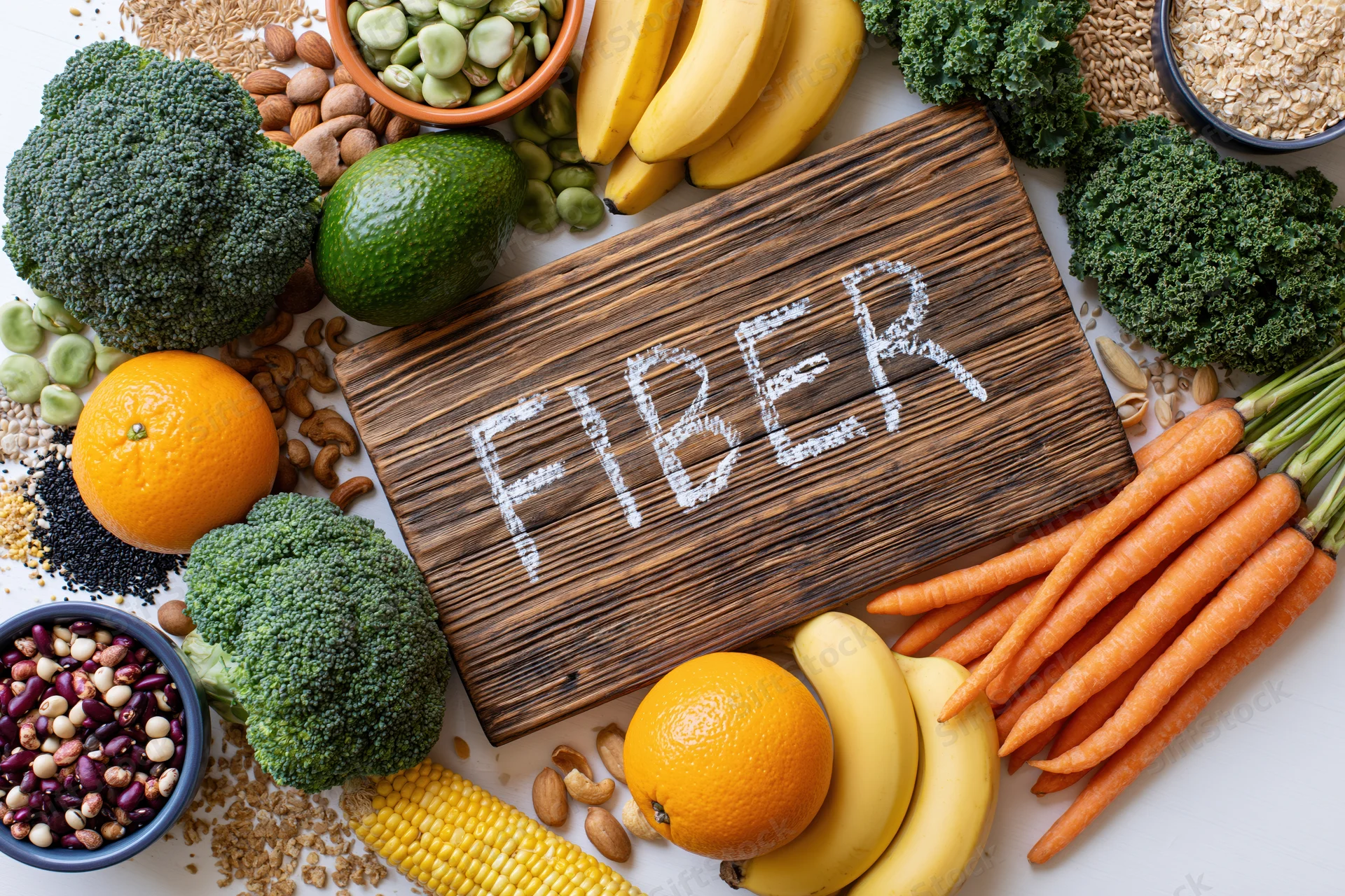 High-fiber foods arranged around a wooden board with fiber written in powder on white surface