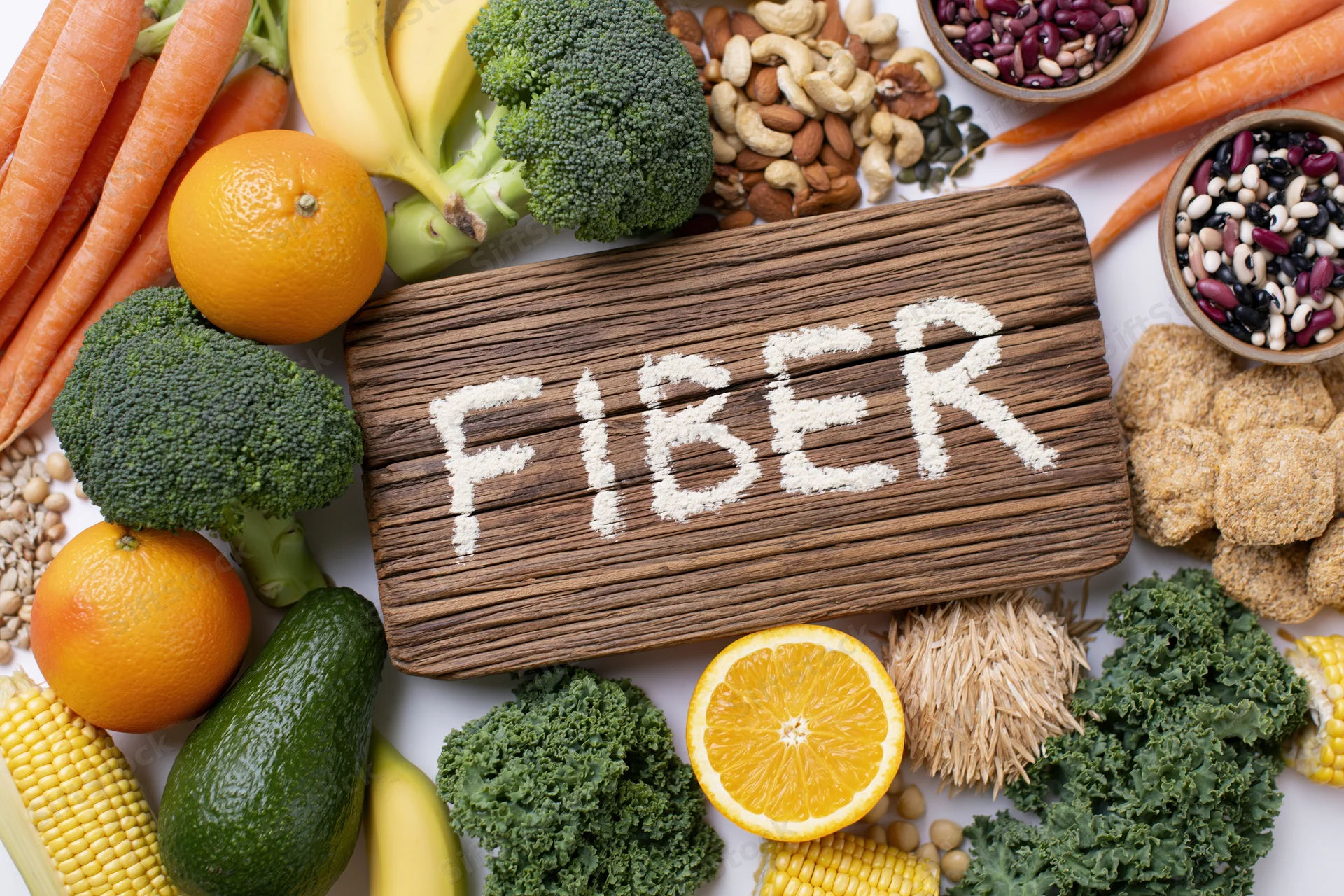 High-fiber foods arranged around a wooden board with fiber written in powder on white surface