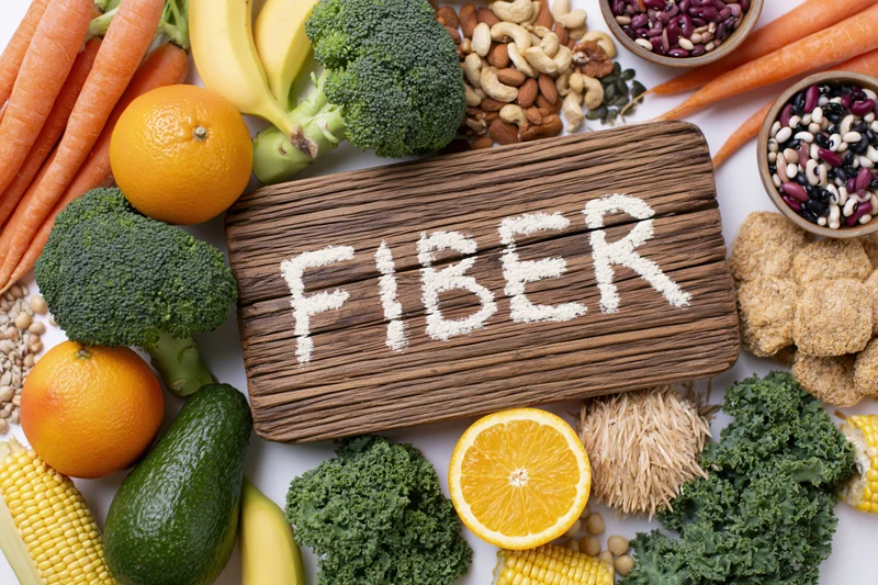 High-fiber foods arranged around a wooden board with fiber written in powder on white surface