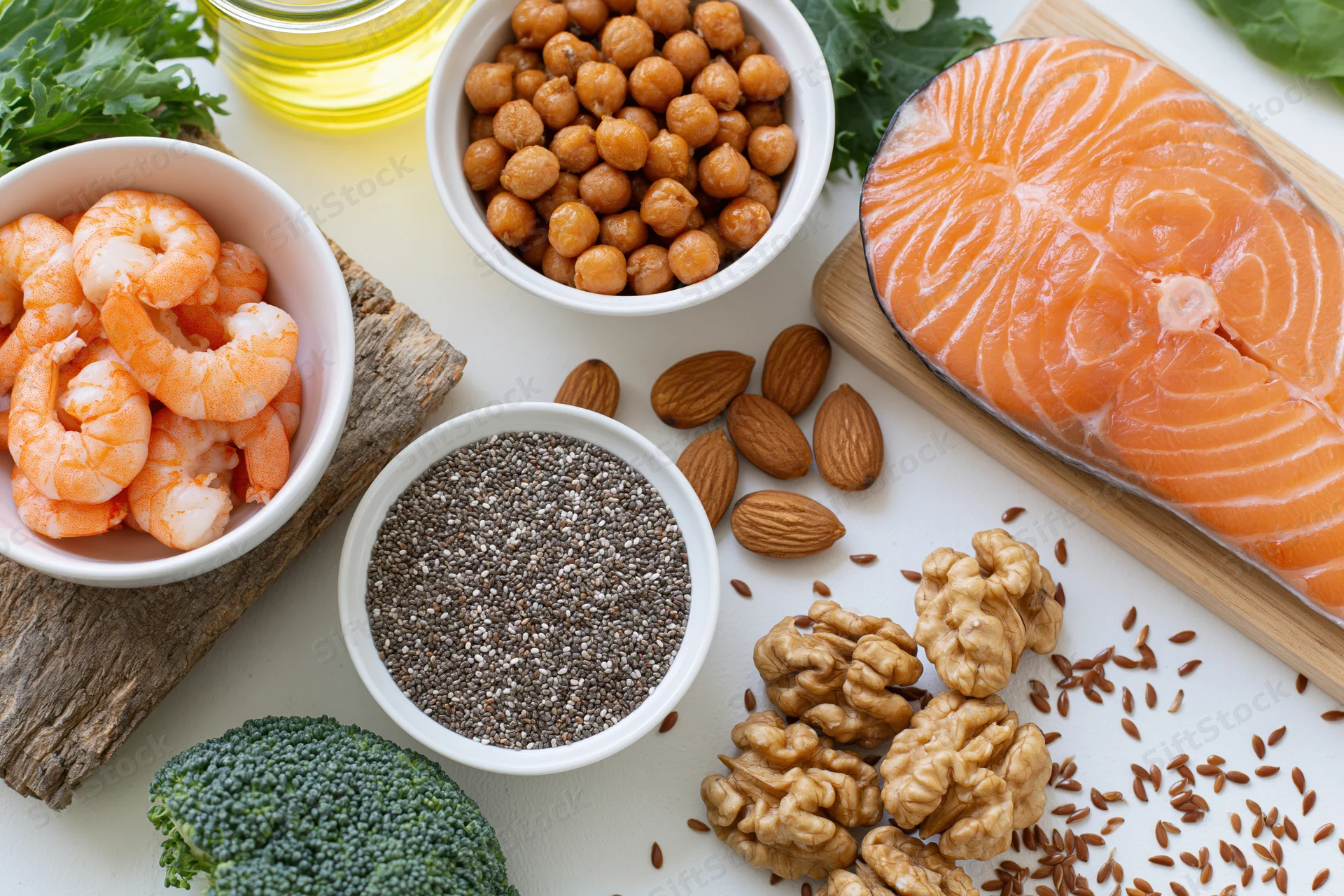 Salmon fillet, shrimp, chia seeds, almonds, walnuts, broccoli, flaxseeds, chickpeas, and oil arranged in bowls and on a white surface in a flat lay composition