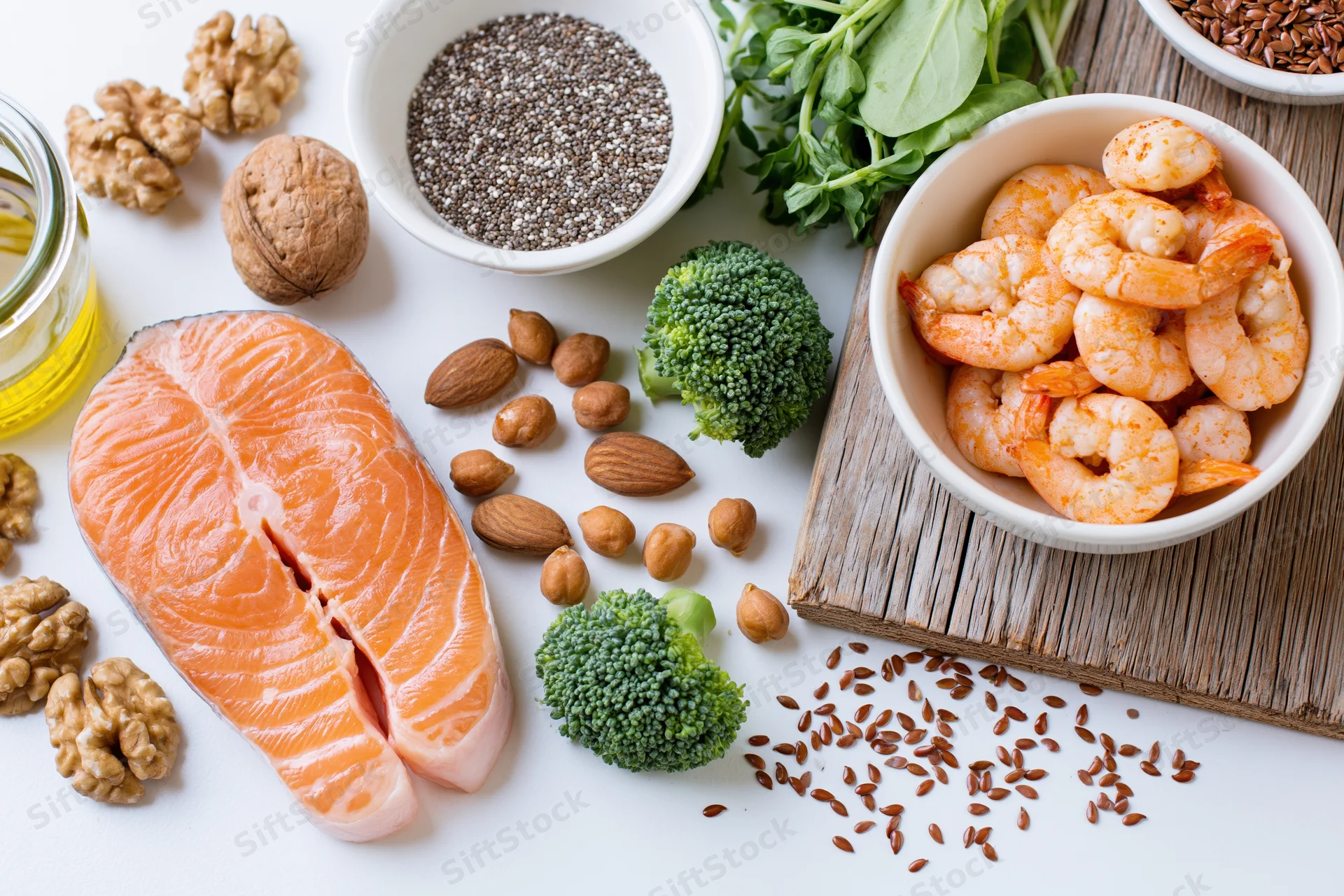 Salmon fillet and shrimp with broccoli, nuts, seeds, and oil arranged on white surface in flat lay composition