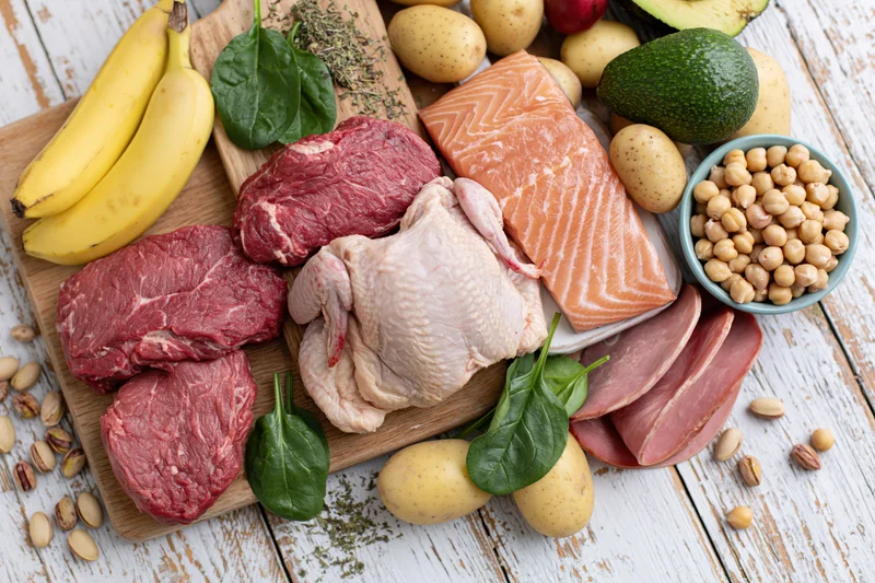 Raw protein-rich foods and fresh produce arranged on a wooden cutting board and white table in a flat lay top view