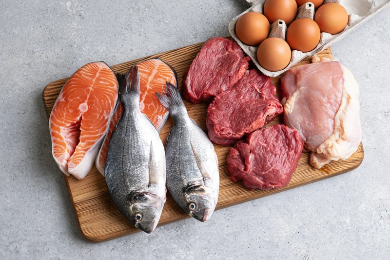 Raw protein foods including salmon, beef, chicken, and eggs arranged on a wooden cutting board on a gray countertop