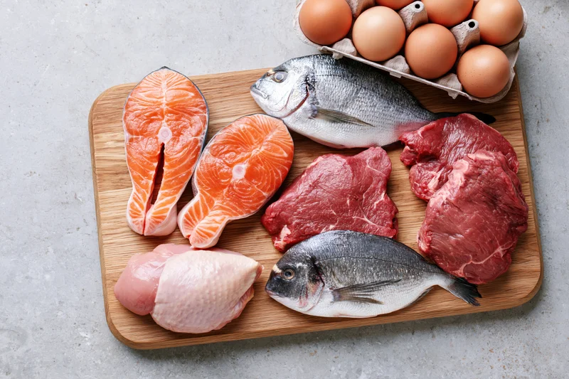 Raw protein foods including salmon, beef, chicken, fish, and eggs arranged on a wooden cutting board in a flat lay top view