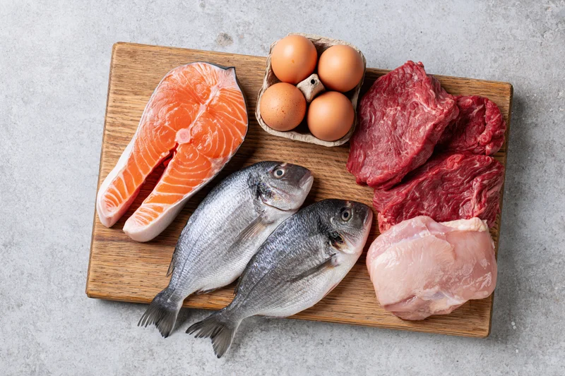 Raw protein foods including salmon, beef, chicken, and eggs arranged on a wooden cutting board in a flat lay overhead view