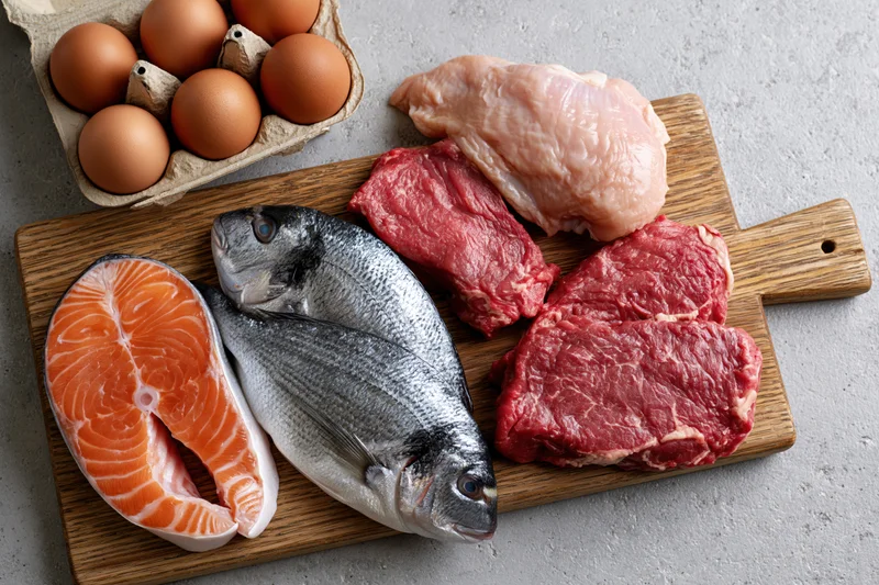 Raw protein foods including salmon, fish, beef, chicken and eggs arranged on a wooden cutting board on a gray countertop in a flat lay overhead view