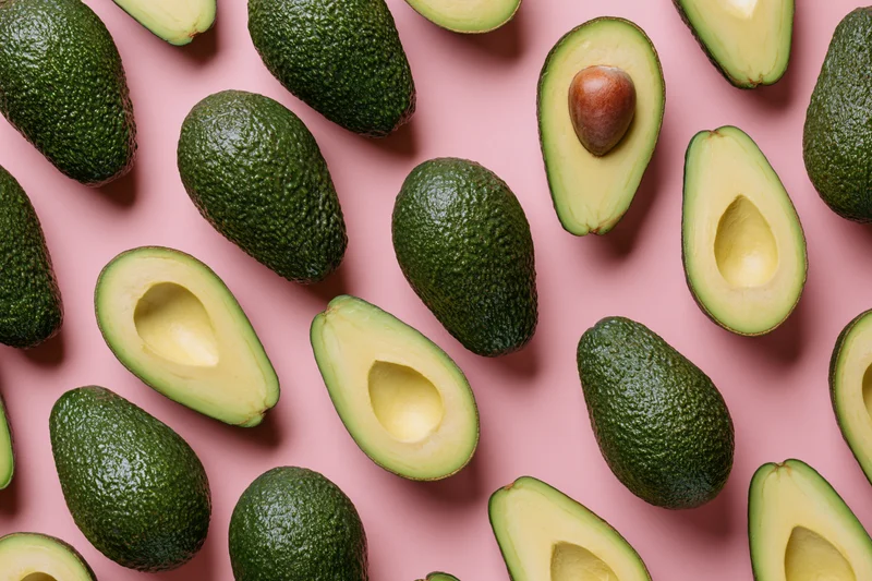 Whole and halved avocados arranged in a repeating pattern on a pink surface