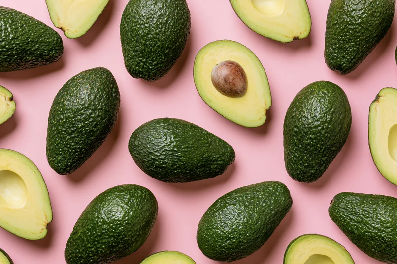 Whole and halved avocados arranged in a repeating pattern on a pink surface