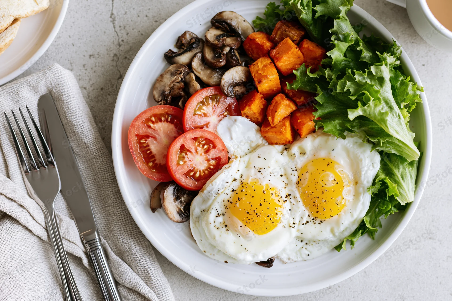 Two fried eggs with tomatoes, mushrooms, sweet potato and lettuce on a white plate with cutlery beside it on a light surface