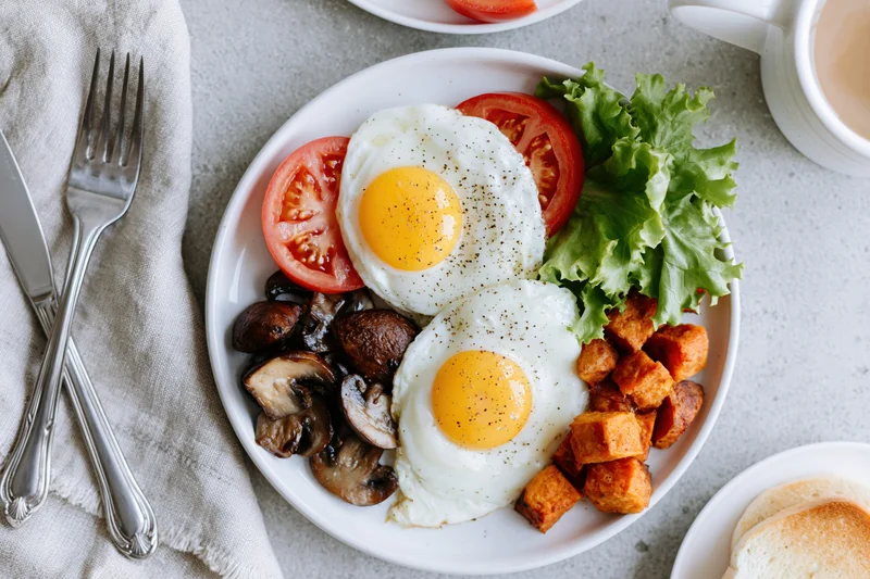 Two fried eggs with tomatoes, lettuce, mushrooms, and tofu on a white plate with cutlery and napkin on a gray surface