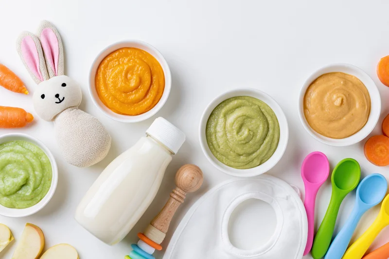 Baby feeding items arranged in flat lay overhead view with purees, bottle, spoons, bib and plush toy on white surface