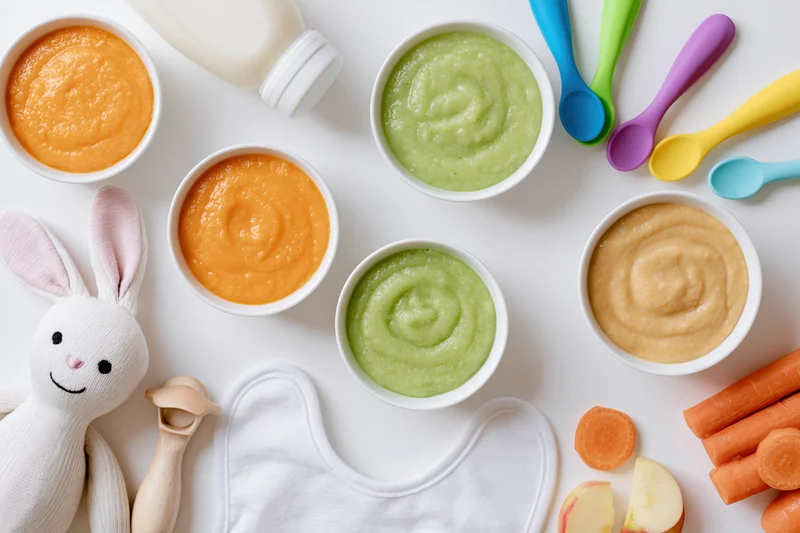 Baby food purees in bowls with spoons, bib, toy, and vegetables arranged on white surface