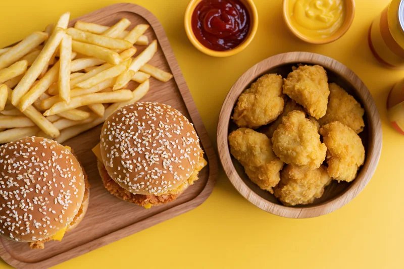 Two burgers with fries and chicken nuggets on a wooden tray with ketchup and mustard on a yellow surface