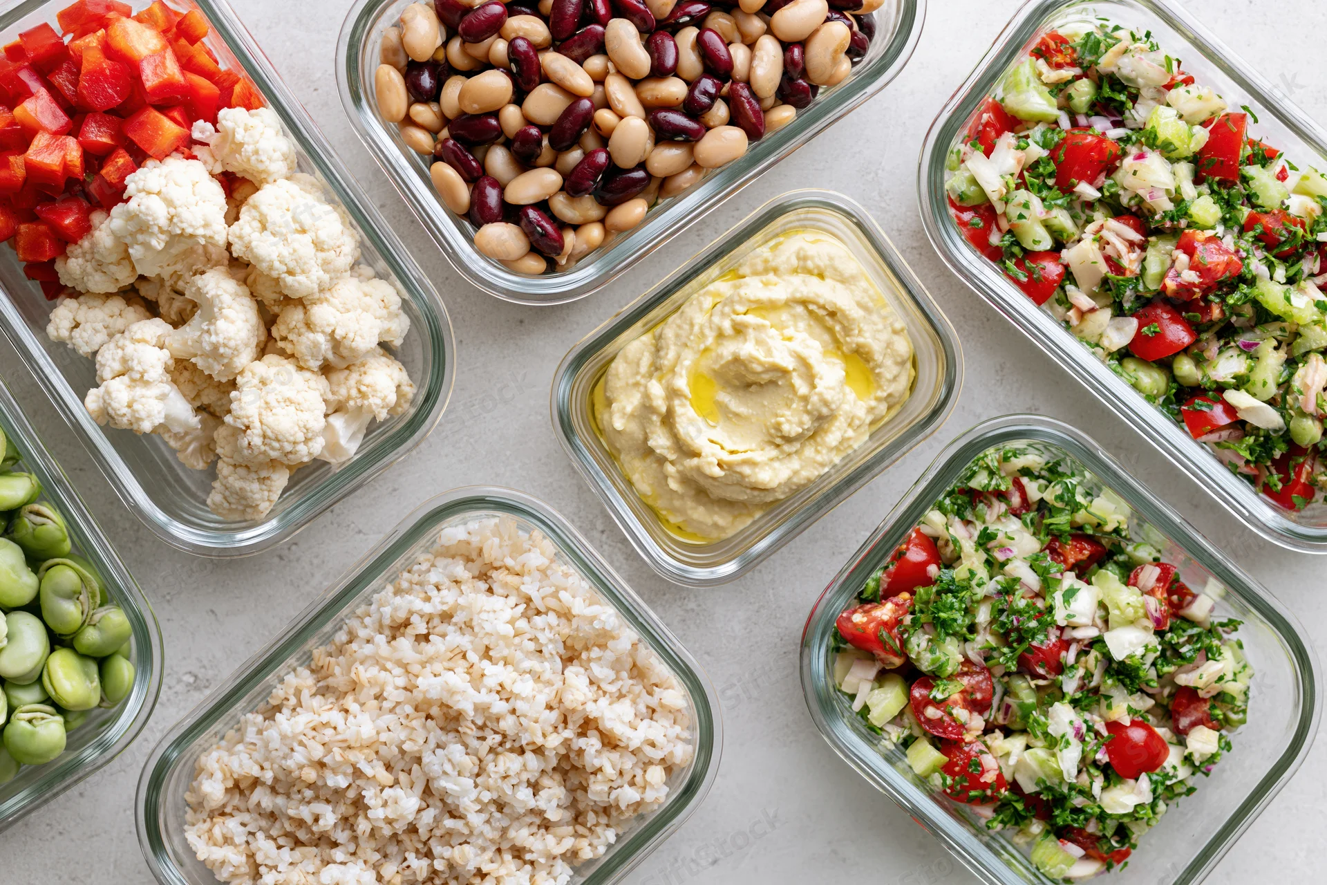 Glass meal prep containers arranged overhead with various healthy foods including vegetables, beans, hummus, rice and salad on a countertop