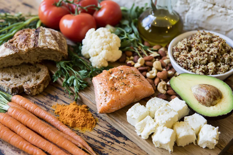 Mediterranean diet ingredients arranged on a wooden table with salmon, avocado, tomatoes, carrots, bread, cheese, nuts, and olive oil