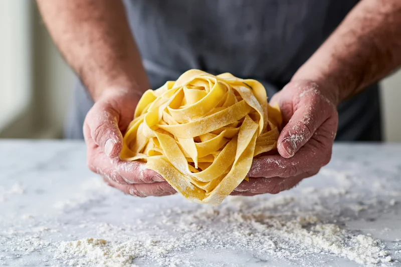 Man’s flour-dusted hands holding a nest of fresh egg pappardelle pasta over a marble countertop