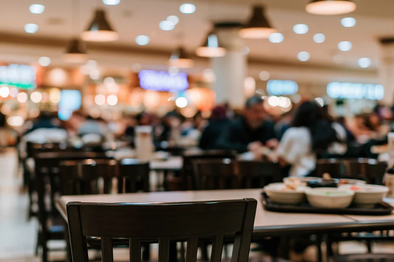 Food court dining area with tables and chairs, people eating in background, low table level view