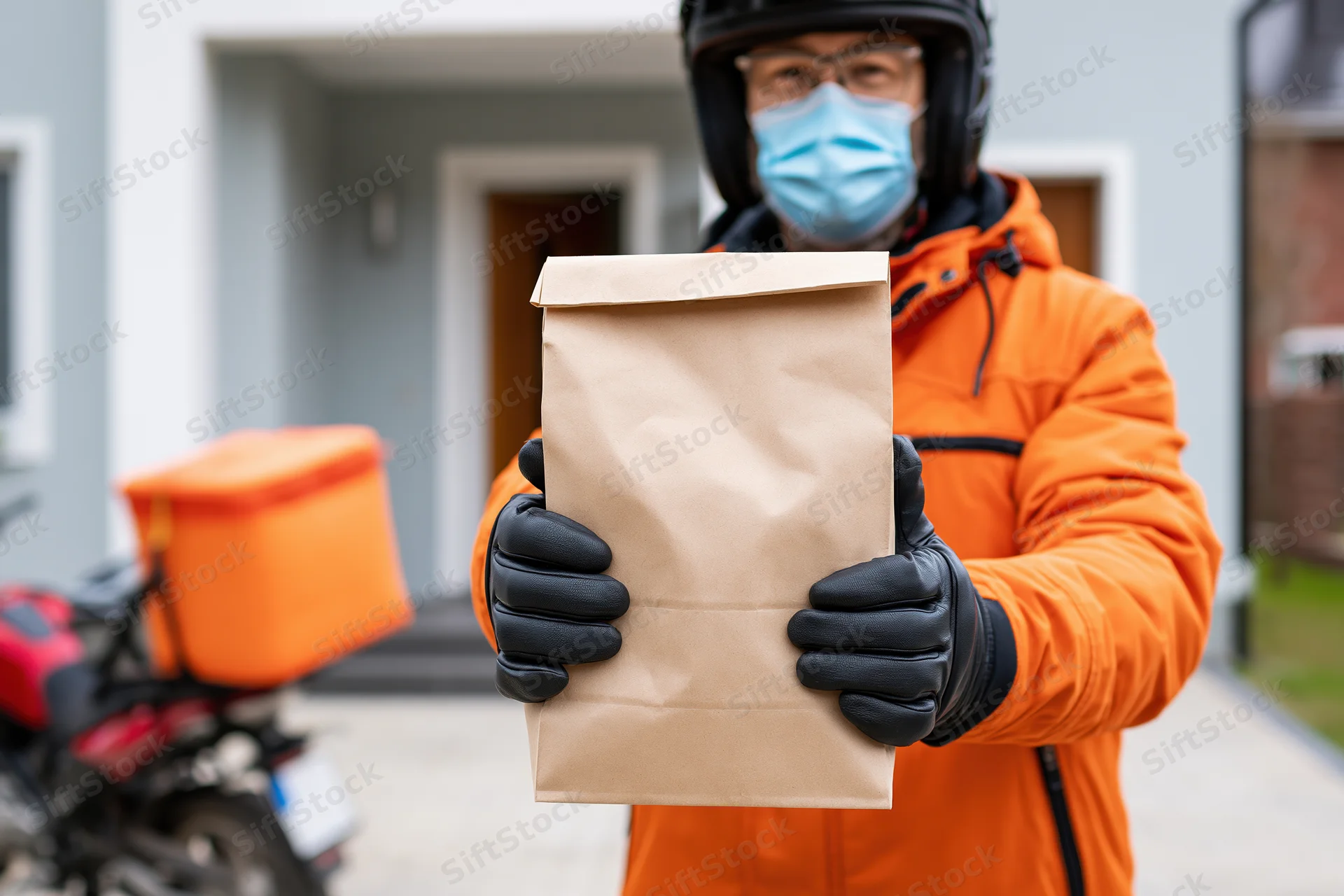 Delivery courier holding food package in residential driveway wearing orange jacket and mask