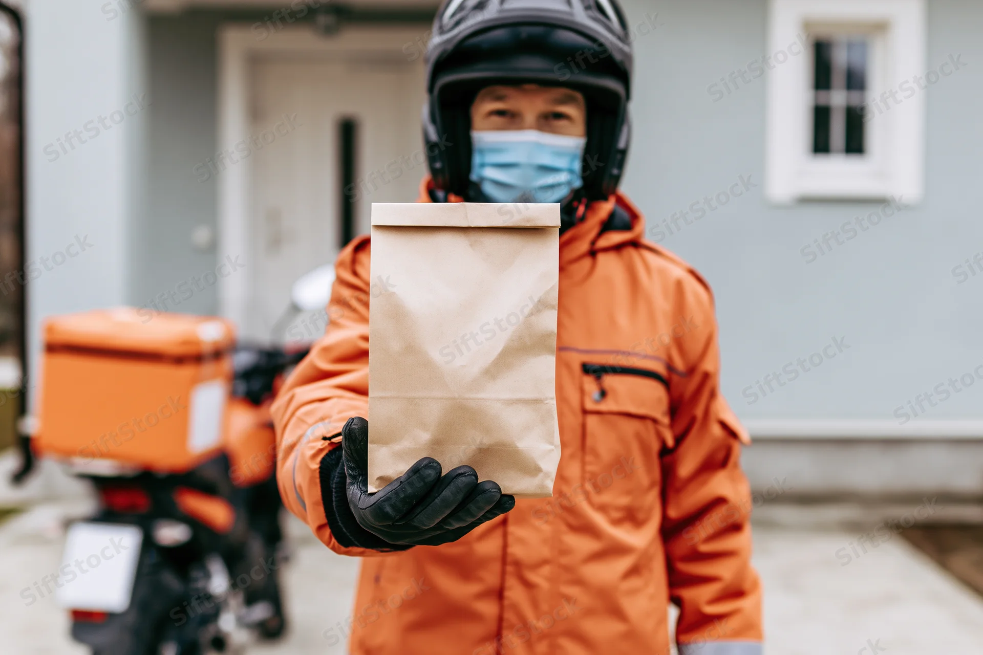 Delivery courier in orange jacket and helmet holding paper food bag in residential driveway
