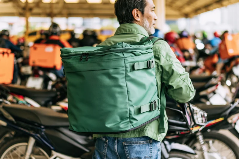 Delivery rider wearing green jacket and carrying large green thermal backpack, standing among motorcycles in urban parking area
