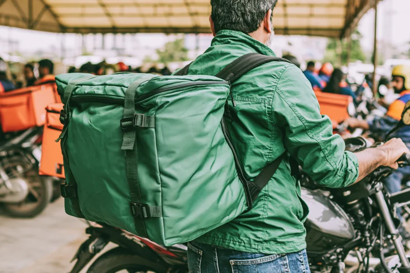 Delivery rider in green jacket with large green backpack on motorcycle in urban setting