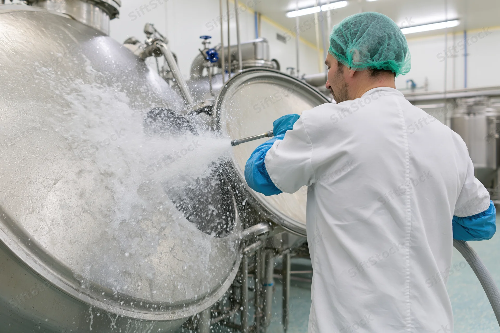 Man in white lab coat and blue gloves spraying water from hose onto large stainless steel tank in food factory