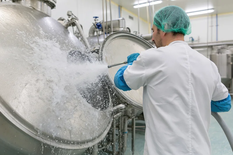 Man in white lab coat and blue gloves spraying water from hose onto large stainless steel tank in food factory