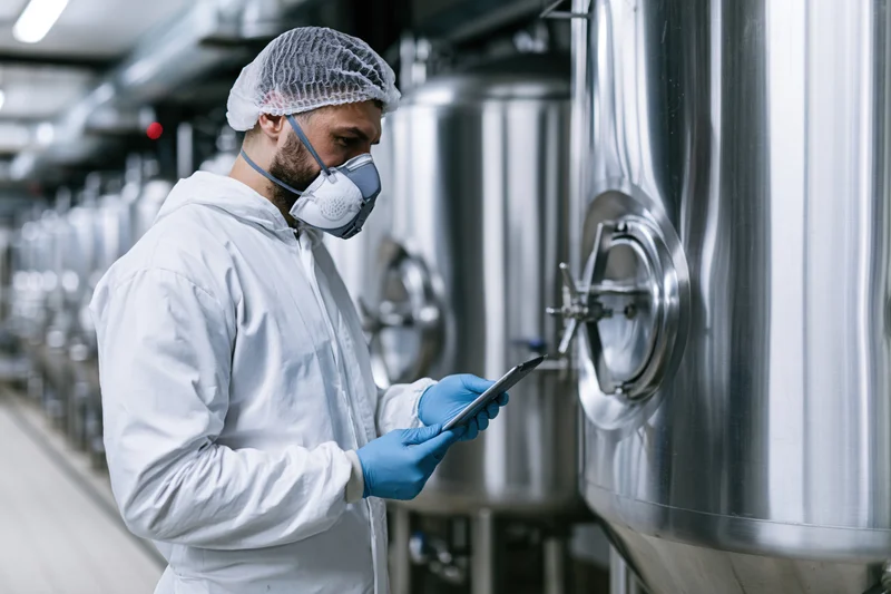 Technician in white coveralls inspecting stainless steel tanks with digital tablet in brewery