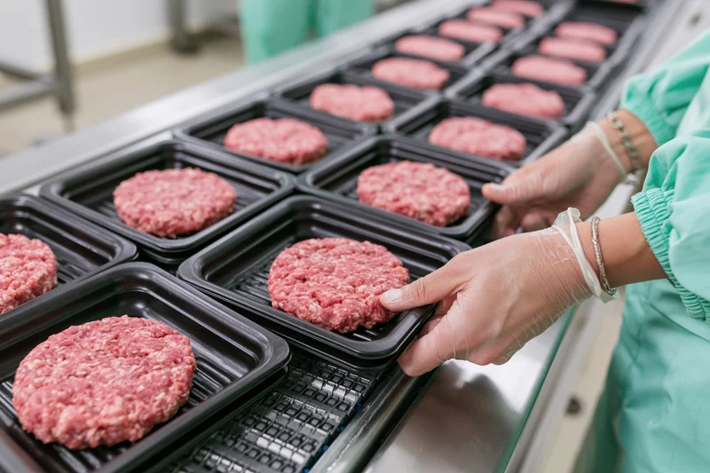 Woman in green coveralls placing raw meat patties into black trays on a conveyor line in a food processing facility