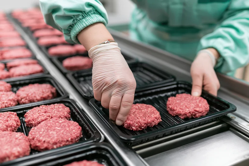 Worker placing raw beef patties into black plastic trays on a conveyor line in a meat processing plant