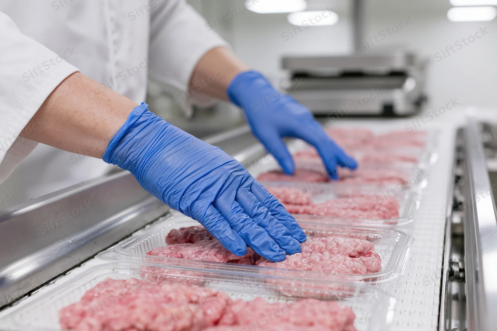 Worker in white lab coat and blue gloves arranging raw ground meat in plastic trays on a conveyor line in a food processing plant