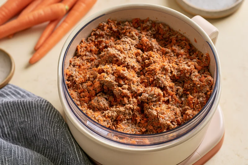 Food processor bowl filled with chopped meat and carrots on a kitchen counter
