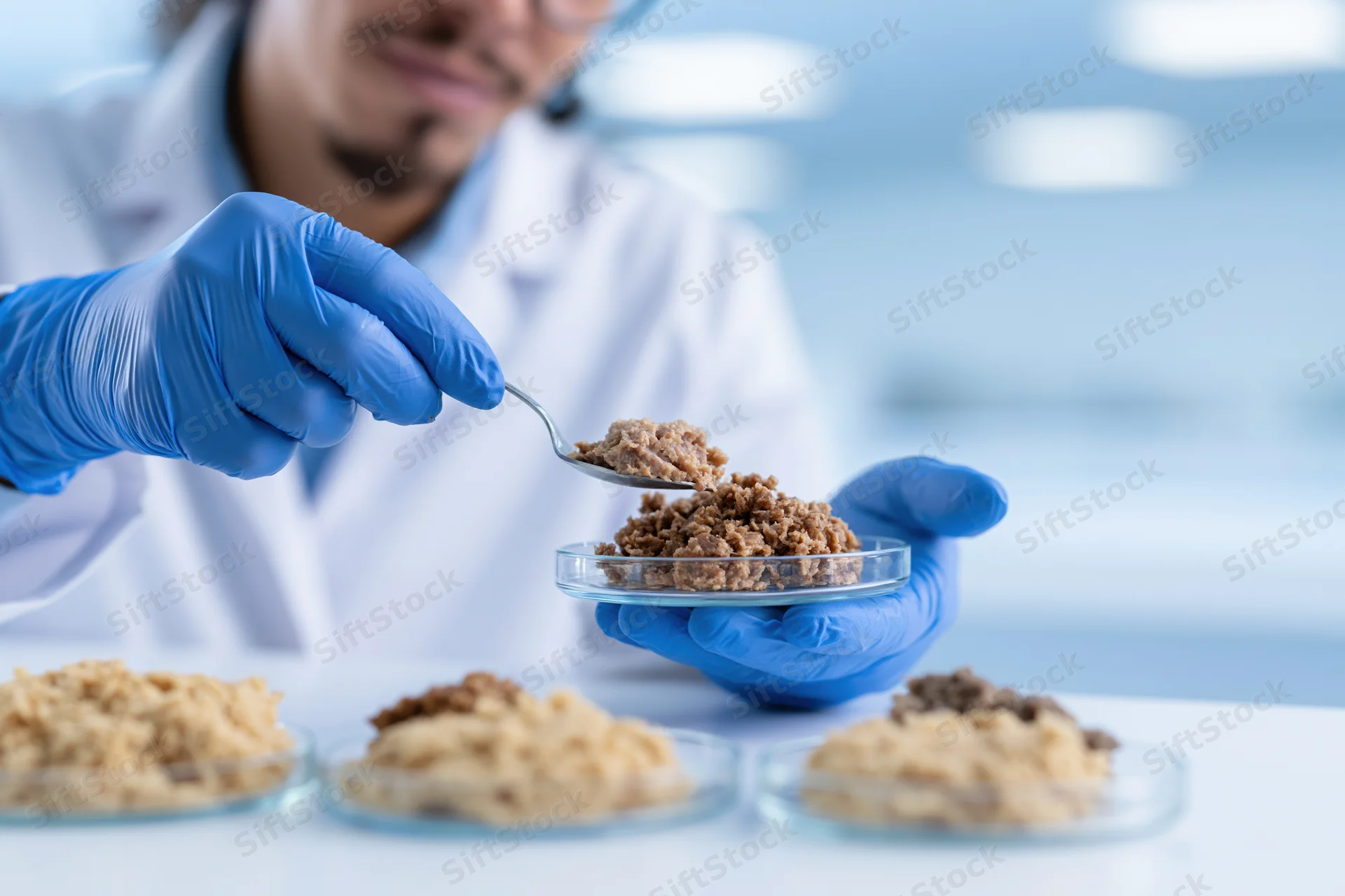 Food scientist in lab coat using spoon to examine meat sample in petri dish in laboratory