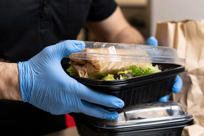 Foodservice worker in blue nitrile gloves holding a black plastic takeout container with salad and sandwich inside