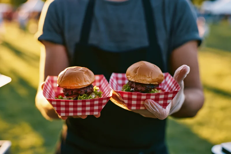 Food vendor holding two burger sliders in red checkered paper baskets outdoors on grass