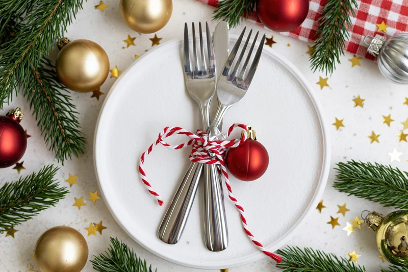 Christmas table setting with cutlery tied with twine and ornaments on a white plate surrounded by pine branches and decorations