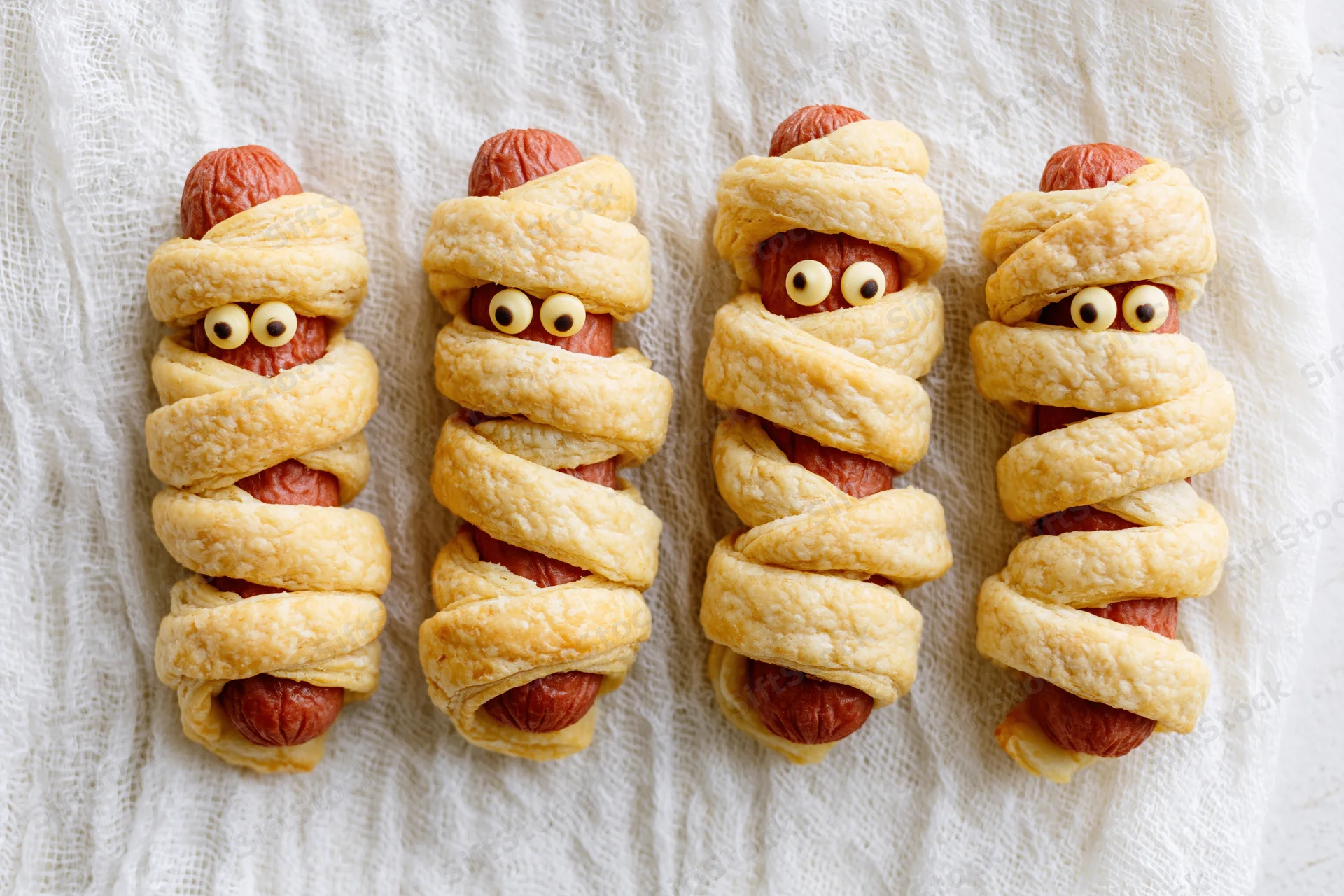 Four mummy-themed hot dogs wrapped in pastry strips with candy eyes on a white cloth surface