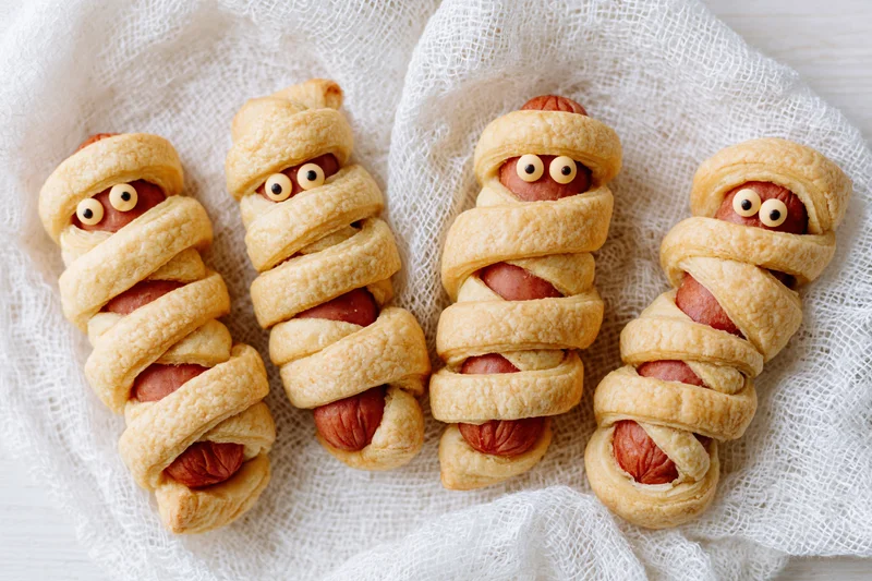 Four mummy-themed hot dogs wrapped in pastry strips with candy eyes on a white mesh cloth