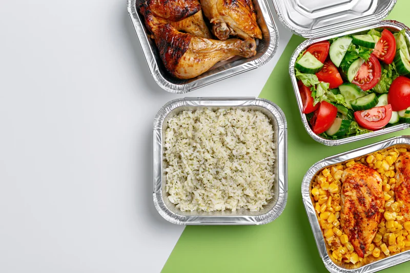 Four aluminum foil takeout containers with chicken, rice, salad and corn arranged on white and green surface