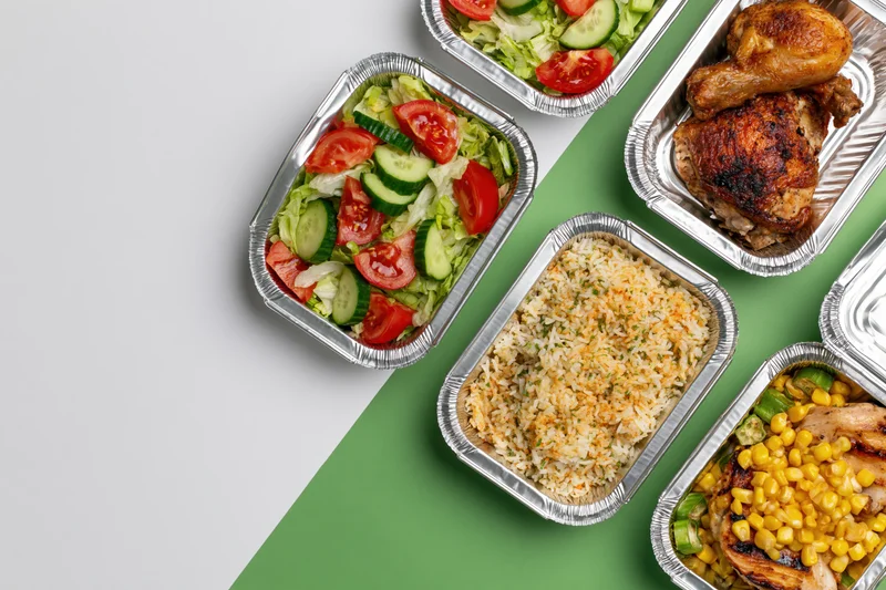 Aluminum foil takeout containers filled with prepared meals arranged on a green and white surface