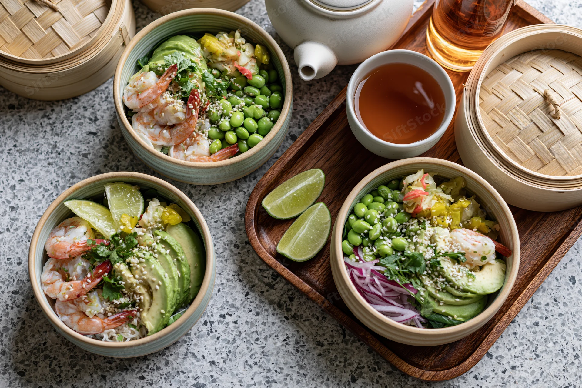 Three poke bowls with shrimp, avocado, edamame and lime on a grey terrazzo surface with bamboo steamers and tea on a wooden tray