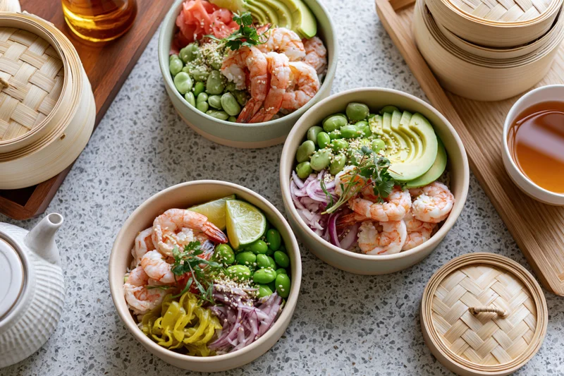 Three ceramic poke bowls with shrimp, edamame, avocado, and garnishes arranged on a grey terrazzo surface with bamboo steamers and tea accessories nearby
