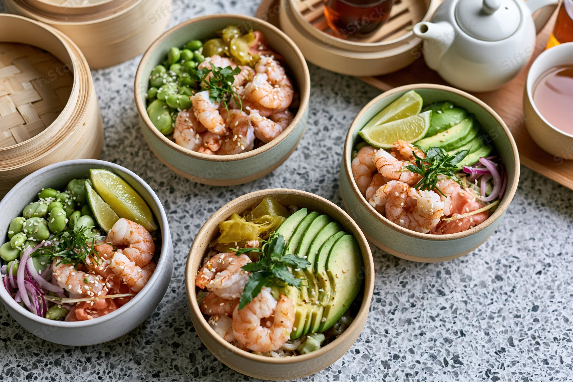 Four ceramic poke bowls with shrimp, avocado, edamame and lime on a grey terrazzo surface with bamboo steamers and tea set