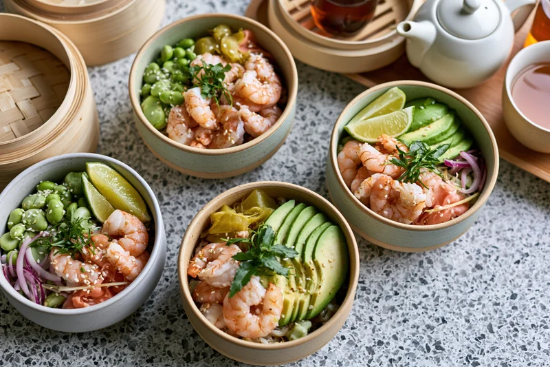 Four ceramic poke bowls with shrimp, avocado, edamame and lime on a grey terrazzo surface with bamboo steamers and tea set