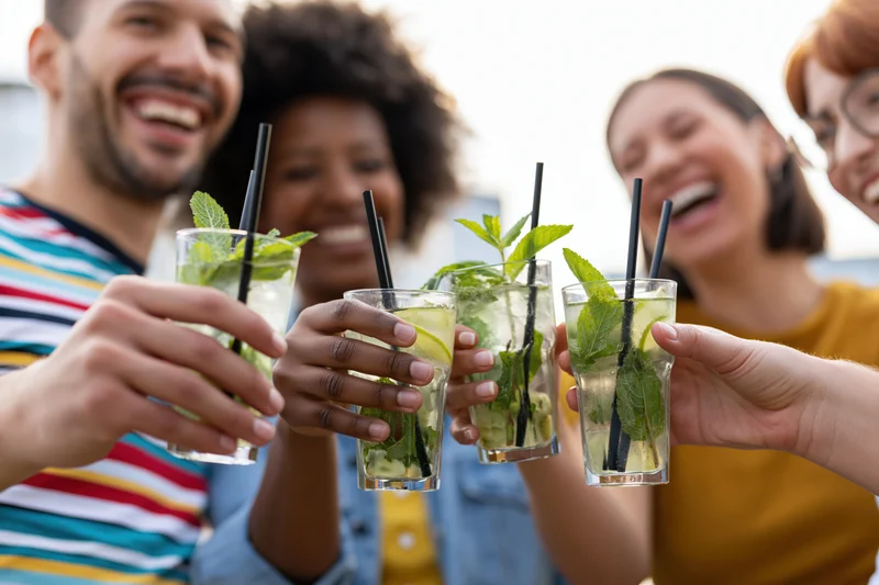 Four diverse young adults laughing and clinking mojito glasses at an outdoor gathering