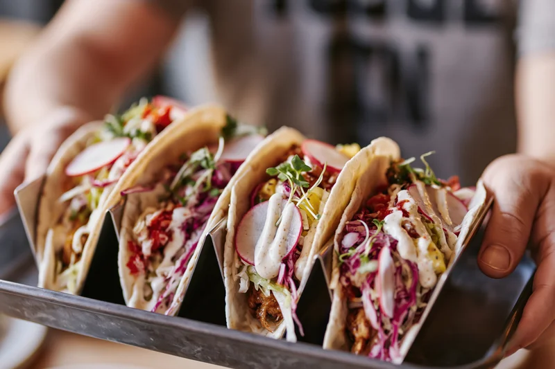 Four gourmet tacos in metal holders on a dark tray held by hands